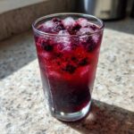 Close-up of a tall, sweating glass filled with vibrant Iced Blackberry Sage Tea, topped with ice cubes and muddled blackberries.