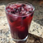 Close-up of a tall, sweating glass filled with vibrant Iced Blackberry Sage Tea, ice cubes, and whole blackberries.