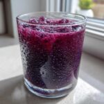 A close-up of a vibrant, deep purple Iced Blueberry Lemonade Slush in a sweating glass, sitting on a bright windowsill.