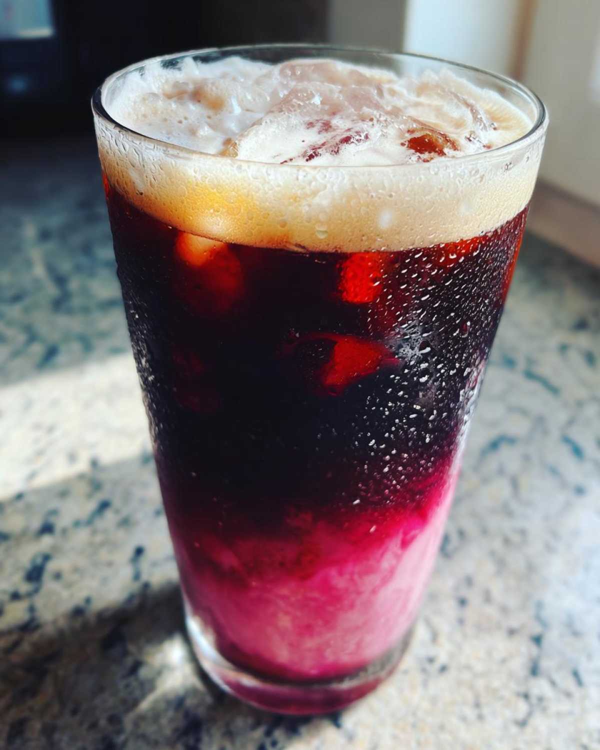 Close-up of a tall glass filled with Iced Cherry Vanilla Cold Brew showing layers of pink syrup, dark coffee, and foam.