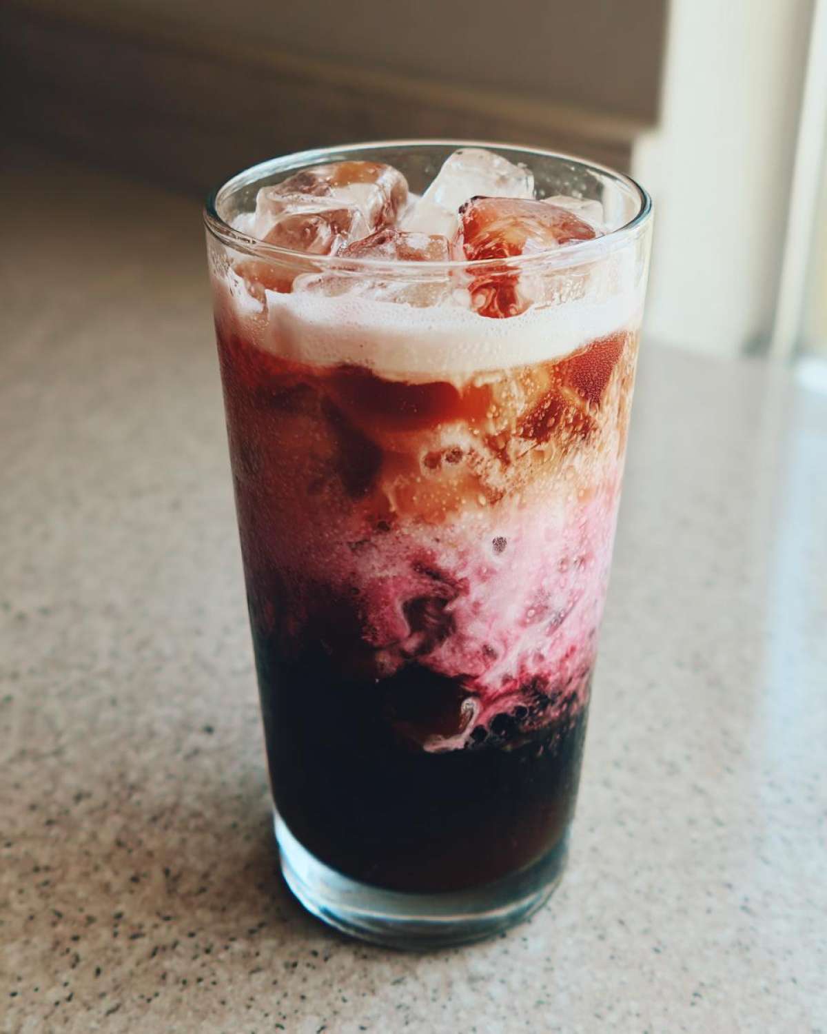Close-up of a tall glass filled with Iced Cherry Vanilla Cold Brew showing dark coffee, pink cherry swirls, and creamy foam topping over ice.