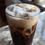Close-up of a tall glass filled with Iced Coconut Mocha with Cold Foam, showing dark coffee and thick white foam.