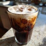 Close-up of a tall glass filled with Iced Coconut Mocha with Cold Foam, showing dark coffee mixing with creamy foam.