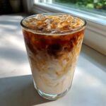 A tall glass showcasing the beautiful swirl of espresso mixing into oat milk for an Iced Honey Vanilla Latte with Oat Milk.