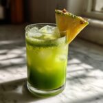 A tall glass filled with a vibrant green Iced Pineapple Matcha Spritz, ice cubes, and garnished with a pineapple wedge.