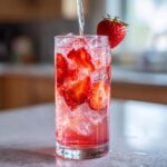 A tall glass filled with ice, sliced strawberries, and pink liquid, as clear liquid is poured in to make Iced Strawberry Hibiscus Tea Lemonade.