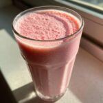 A tall glass filled with a frothy, pink Iced Vanilla Strawberry Protein Shake, glistening with condensation.