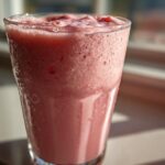 Close-up of a tall, frosty glass filled with a pink, frothy Iced Vanilla Strawberry Protein Shake, condensation visible on the glass.