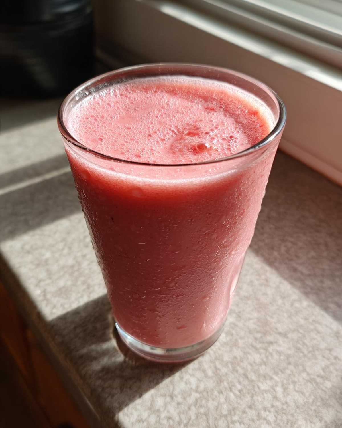 A tall glass filled with a cold, pink Iced Vanilla Strawberry Protein Shake, showing condensation on the outside.