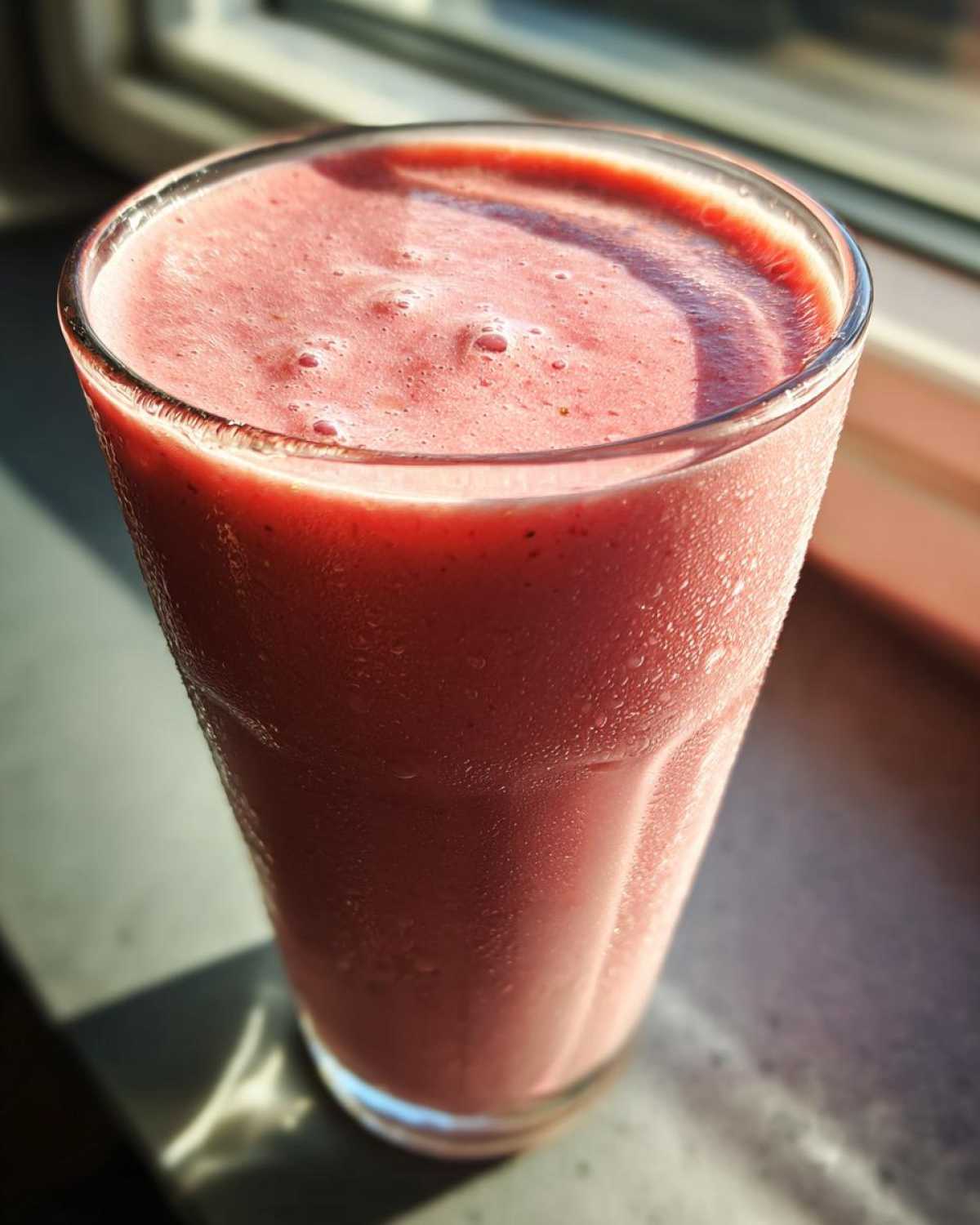 Close-up of a tall glass filled with a frothy, pink Iced Vanilla Strawberry Protein Shake, showing condensation on the glass.