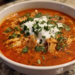 Close-up of a white bowl filled with rich, red Instant Pot Creamy Chicken Enchilada Soup, topped with sour cream and cilantro.