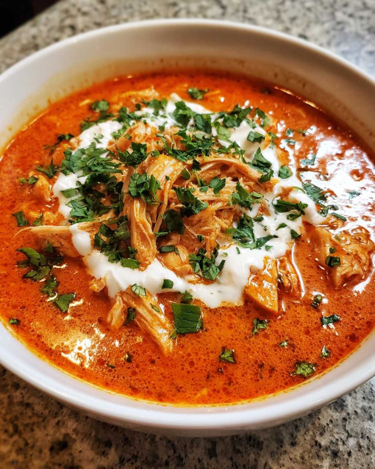 Close-up of a white bowl filled with vibrant orange Instant Pot Creamy Chicken Enchilada Soup, topped with shredded chicken, sour cream, and fresh parsley.
