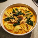 A close-up bowl of rich, orange-hued Instant Pot Creamy Chicken Tortellini Soup topped with shredded chicken, wilted spinach, and red pepper flakes.