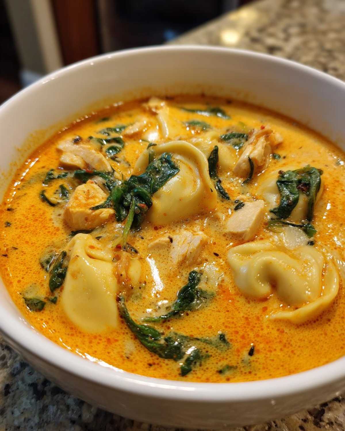Close-up of a white bowl filled with Instant Pot Creamy Chicken Tortellini Soup, showing tortellini, chicken chunks, and spinach in a rich orange broth.