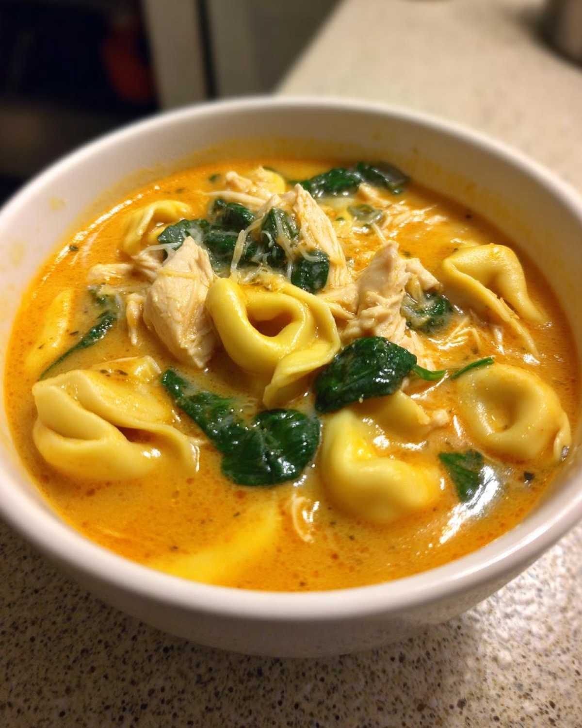 Close-up of a white bowl filled with rich, orange-hued Instant Pot Creamy Chicken Tortellini Soup, featuring tortellini, shredded chicken, and spinach.