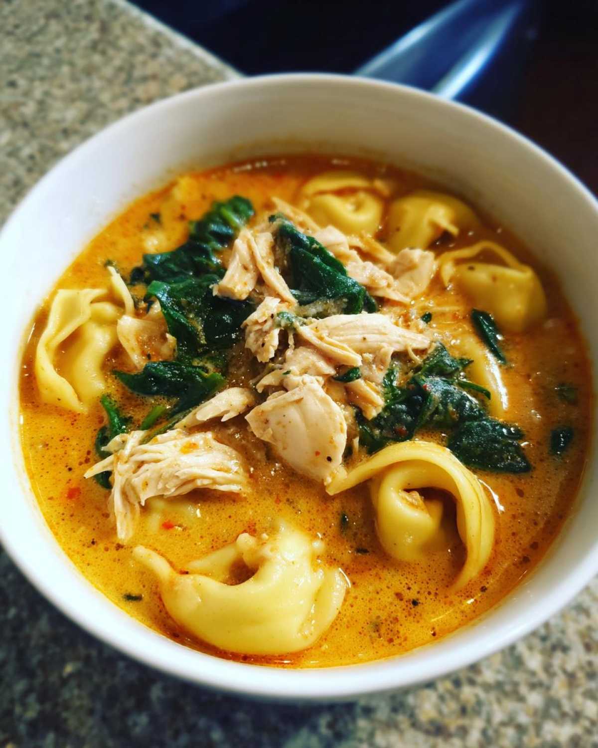 Close-up of a white bowl filled with Instant Pot Creamy Chicken Tortellini Soup, featuring shredded chicken, spinach, and tortellini.