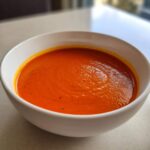 Close-up of a white bowl filled with vibrant orange Instant Pot Creamy Red Pepper Tomato Soup.
