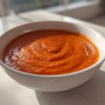 Close-up of a white bowl filled with vibrant orange Instant Pot Creamy Red Pepper Tomato Soup, garnished with pepper flecks.