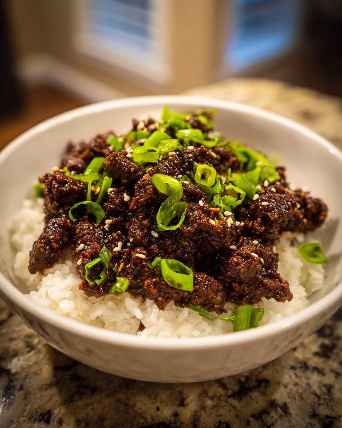 Close-up of a white bowl filled with white rice topped with saucy Instant Pot Korean Beef and garnished with green onions and sesame seeds.