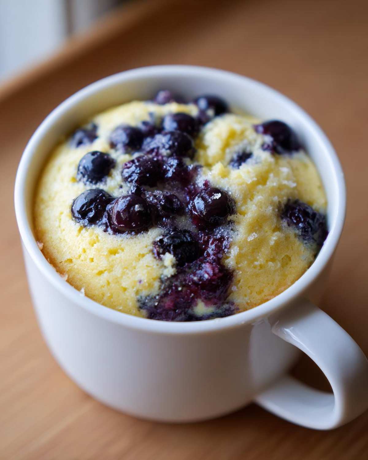 Close-up of a freshly baked Keto Blueberry Lemon Mug Cake in a white mug, topped with juicy blueberries.