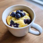 Close-up of a single serving Keto Blueberry Lemon Mug Cake baked in a white mug, topped with fresh blueberries and powdered sweetener.