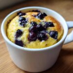 Close-up of a golden Keto Blueberry Lemon Mug Cake baked in a white mug, topped with juicy blueberries.
