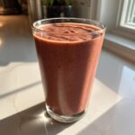 A tall glass filled with a thick, reddish-brown Keto Chocolate Strawberry Smoothie sitting on a white marble counter.