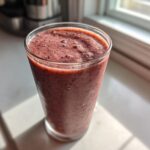 A tall glass filled with a thick, dark red Keto Chocolate Strawberry Smoothie sitting on a sunlit windowsill.