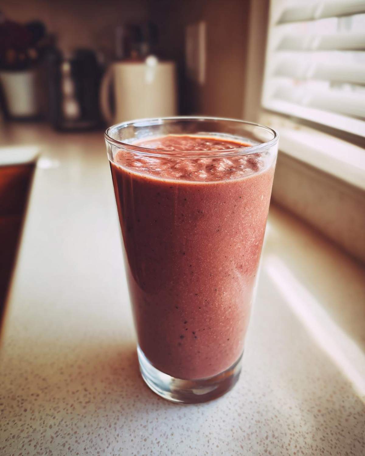 A tall glass filled with a thick, reddish-brown Keto Chocolate Strawberry Smoothie sitting on a light countertop near a window.