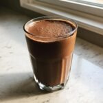 A tall glass filled with a thick, dark brown Keto Creamy Chocolate Peanut Butter Shake, sitting on a white countertop near a window.