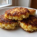 A stack of four golden-brown, crispy Keto Crispy Cauliflower Hash Browns on a white plate.