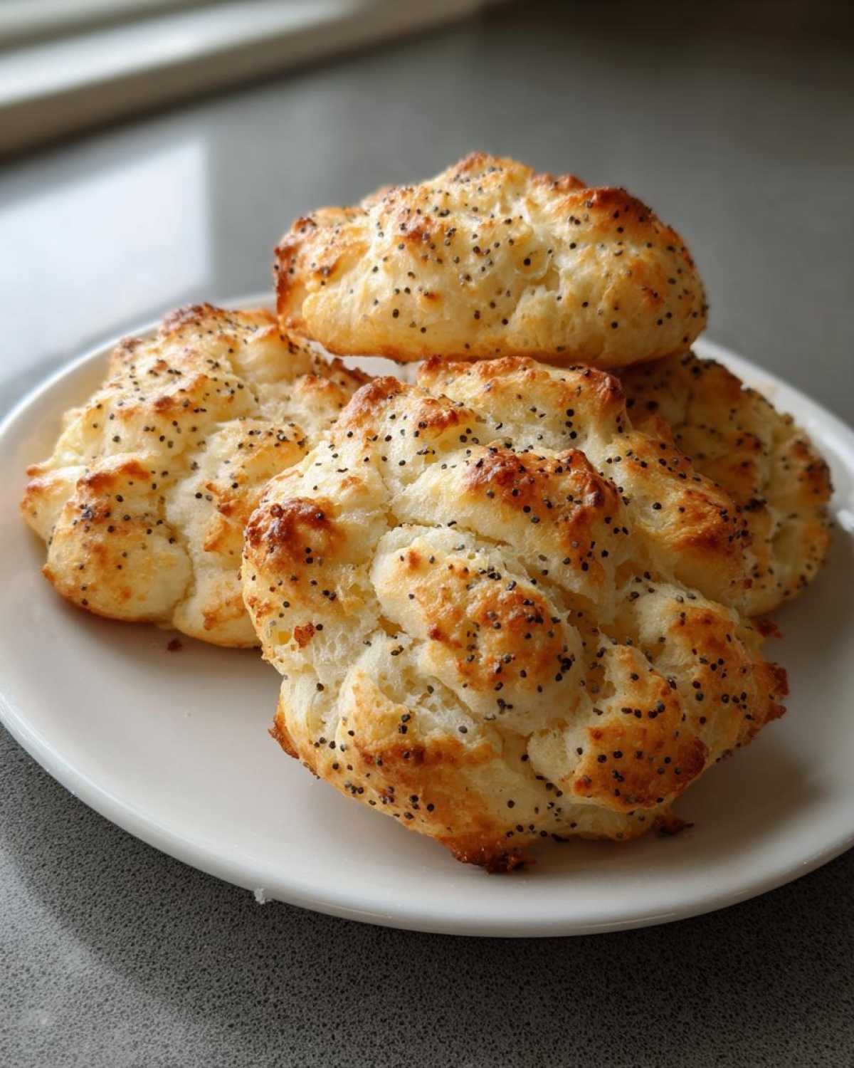Four golden brown Keto Everything Bagel Cloud Bread puffs topped with poppy seeds stacked on a white plate.