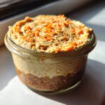 A single serving of No-Bake Carrot Cake Cheesecake Cups layered in a small glass jar, topped with shredded carrots.