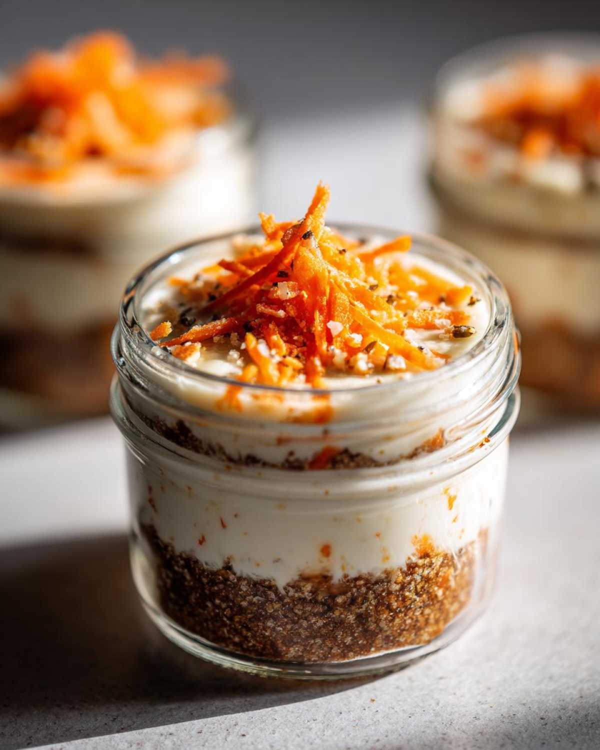 Close-up of a No-Bake Carrot Cake Cheesecake Cups layered in a small glass jar, topped with shredded carrots.