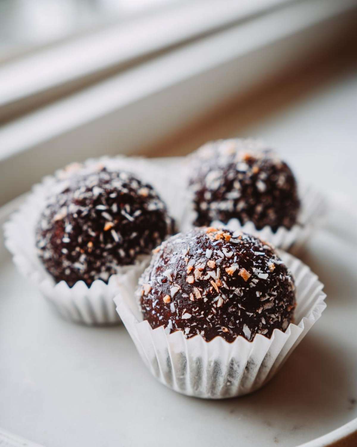 Three rich, dark No-Bake Chocolate Coconut Protein Cups rolled in shredded coconut, sitting in white paper liners.