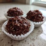 Three No-Bake Chocolate Coconut Protein Cups sitting in white paper liners on a speckled countertop.