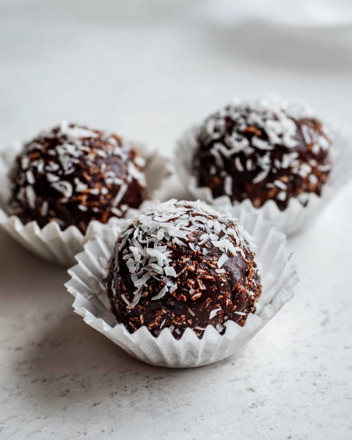 Three No-Bake Chocolate Coconut Protein Cups coated in shredded coconut, resting in white paper liners.