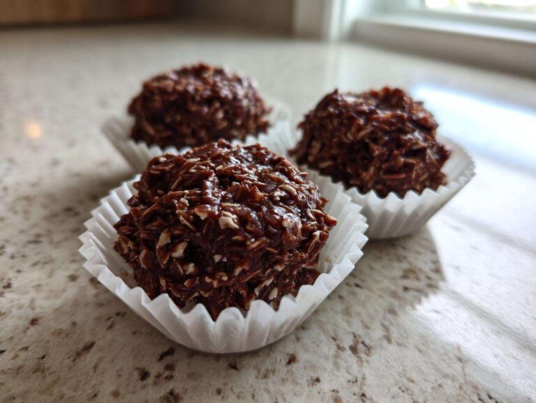 Three No-Bake Chocolate Coconut Protein Cups sitting in white paper liners on a speckled countertop.