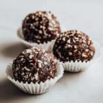 Three rich, dark No-Bake Chocolate Coconut Protein Cups rolled in shredded coconut on a white surface.
