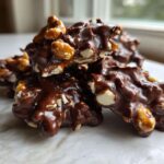 A stack of rich, glossy No-Bake Chocolate Covered Pretzel Clusters featuring visible nuts and pretzel pieces.