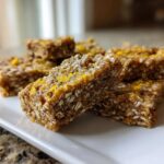 Close-up of several No-Bake Lemon Almond Date Bars stacked on a white rectangular plate, topped with yellow zest.