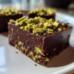 Close-up of rich, dark No-Bake Pistachio Chocolate Fudge Squares topped with chopped green pistachios on a white plate.