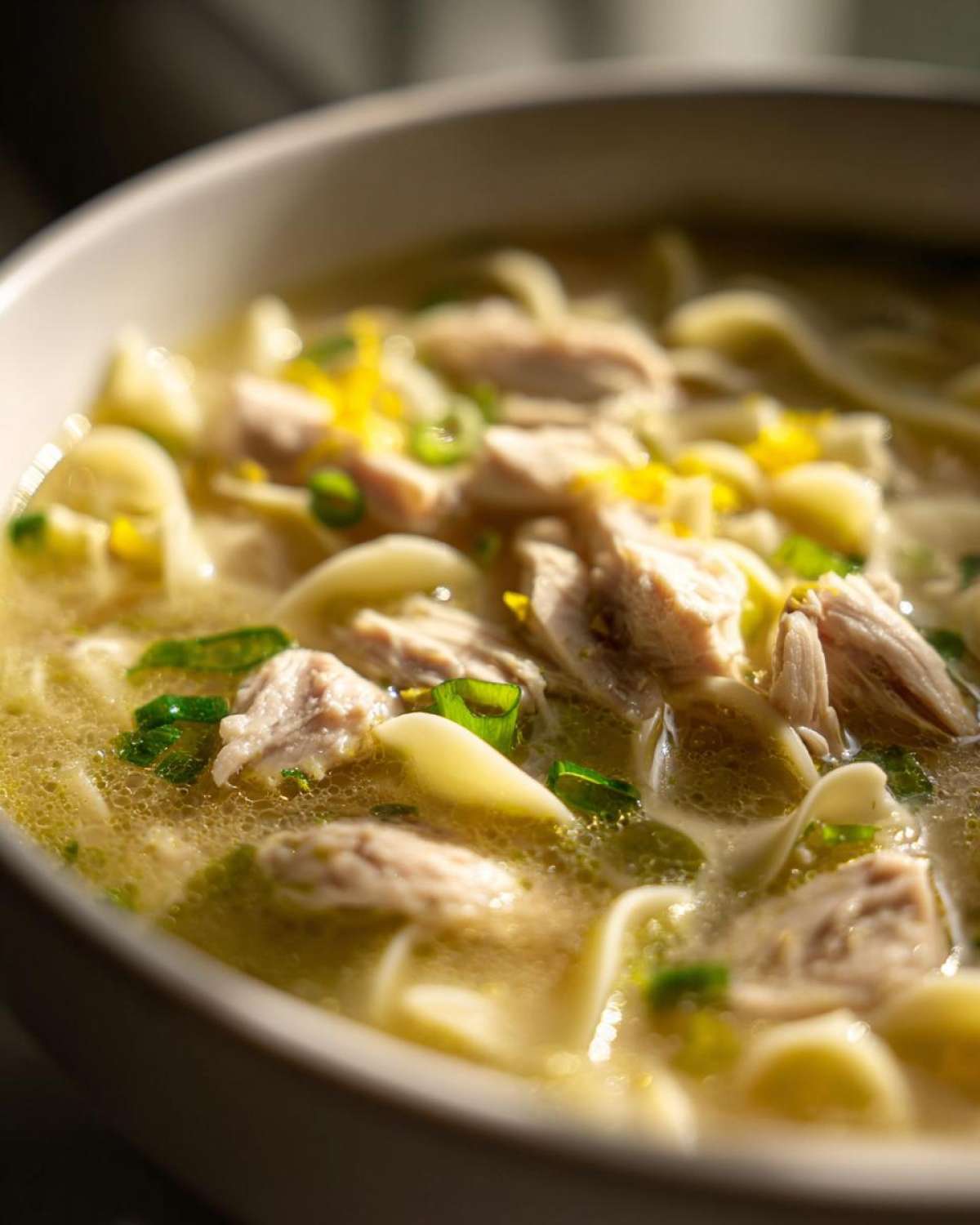 Close-up of chunks of chicken and egg noodles in a broth, garnished with lemon zest and green onions in a bowl of One-Pot Creamy Lemon Chicken Noodle Soup.