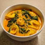 A close-up of a white bowl filled with rich, orange-hued One-Pot Creamy Tortellini Minestrone soup featuring tortellini and wilted spinach.