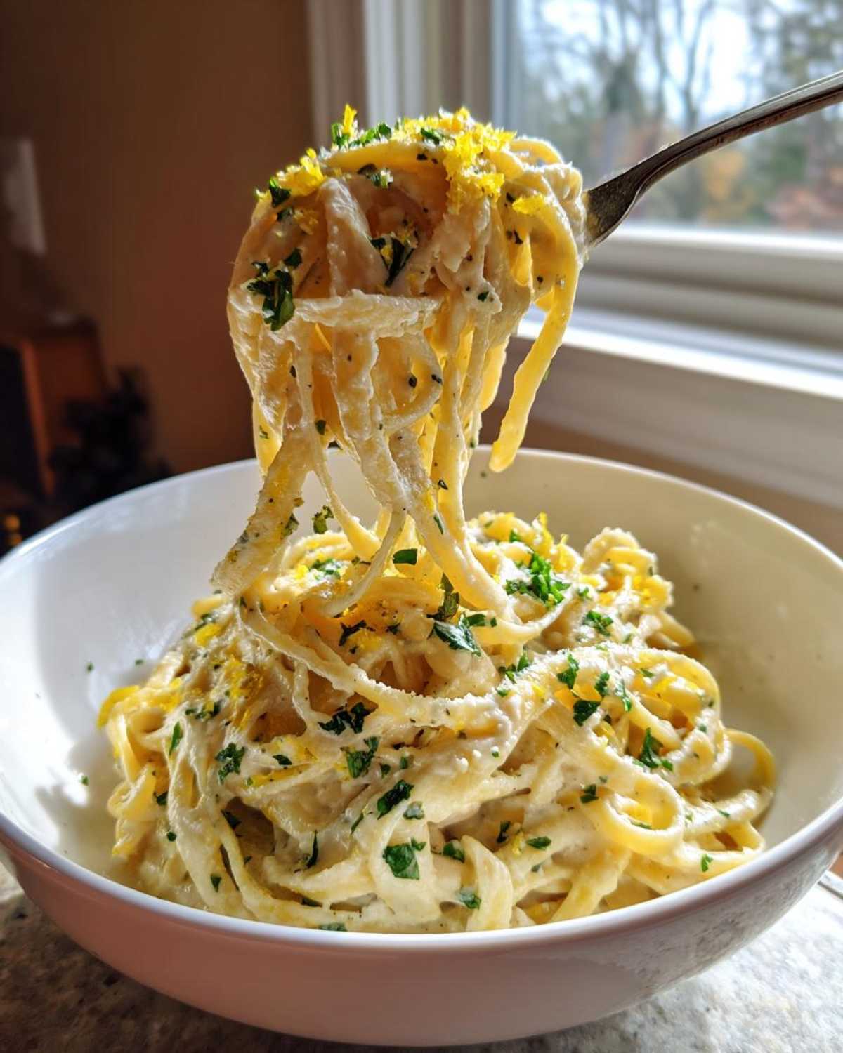 A fork lifts creamy strands of One-Pot Creamy White Bean Lemon Pasta, garnished with lemon zest and parsley, from a white bowl.