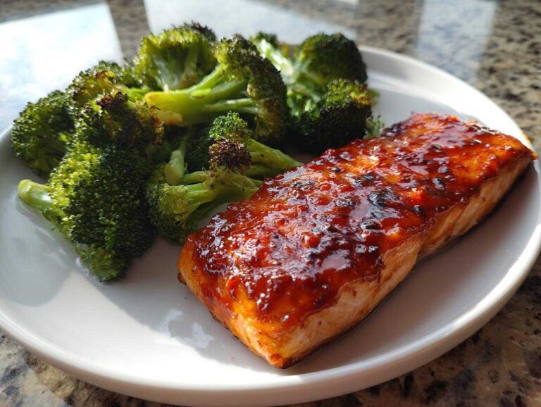 A glistening fillet of Sheet Pan Honey Harissa Salmon served next to bright green roasted broccoli florets on a white plate.