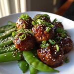 Close-up of glazed Sheet Pan Sticky Sesame Meatballs served over bright green snap peas, garnished with sesame seeds and scallions.