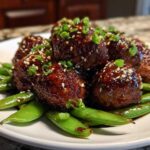 Close-up of glazed Sheet Pan Sticky Sesame Meatballs with Snap Peas garnished with sesame seeds and green onions.