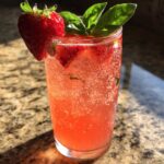 Close-up of a tall glass filled with vibrant Sparkling Strawberry Basil Lemonade, garnished with a fresh strawberry and basil leaves.