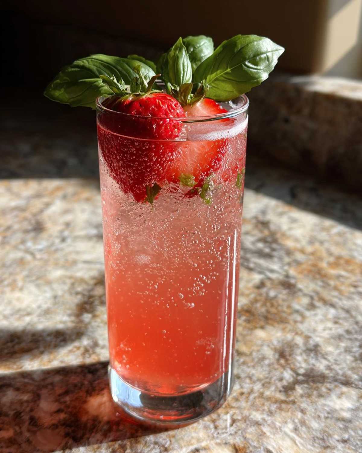 A tall glass filled with vibrant pink Sparkling Strawberry Basil Lemonade, garnished with fresh strawberries and basil leaves.
